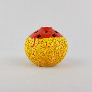 May include: A round, ceramic flower frog with a textured yellow body and a red top. The flower frog has several holes for arranging stems. The yellow surface has a cracked, aged appearance, adding a unique aesthetic to the piece.