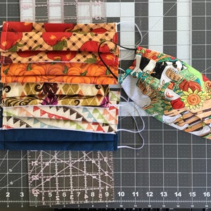May include: Five fabric face masks with different patterns. The masks are layered and sewn together. The patterns include pumpkins, fall leaves, and a farmhouse scene. The masks are on a gray cutting mat with a ruler.