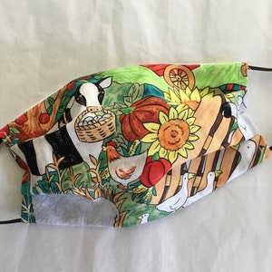 May include: A colorful fabric face mask with a farm scene featuring a cow, chickens, ducks, pumpkins, sunflowers, and a wheelbarrow. The mask is green, yellow, red, black, and white.