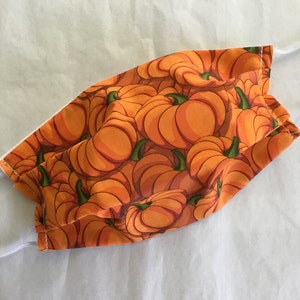 May include: A reusable face mask with a bright orange and green pumpkin print. The mask is made of fabric and has a white elastic band.