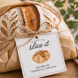 May include: A loaf of artisan bread wrapped in brown paper, tied with twine, and a tag that reads "Any way you slice it, we are so thankful for you!" The bread has a decorative leaf pattern.