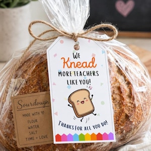 Könnte beinhalten: Ein Brotlaib Sauerteigbrot, verpackt in durchsichtigem Zellophan, mit Bindfaden gebunden, und ein Geschenkanhänger mit der Aufschrift "We Knead More Teachers Like You!" mit einem Cartoon-Brotscheibe. Ein kleines Etikett lautet "Sauerteig".