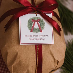 May include: A Christmas gift wrapped in brown paper, secured with a burgundy ribbon and twine. A tag features a watercolour wreath and the words "Freshly Baked With Love. Merry Christmas". The gift is on a red and white checked surface.