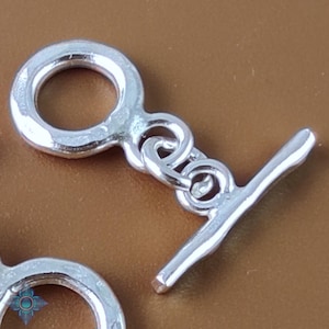 Könnte beinhalten: Nahaufnahme eines silberfarbenen Knebelverschlusses. Der Verschluss hat einen kreisförmigen Ring und einen T-Stab, die durch Schlaufen verbunden sind. Das Metall hat eine glänzende, reflektierende Oberfläche. Der Hintergrund ist ein warmes Braun.