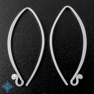 Puede incluir: Un par de pendientes plateados con forma de lágrima alargada. Los pendientes tienen un diseño liso y curvado con un pequeño remolino decorativo en la parte inferior. El fondo es una superficie oscura y texturizada.