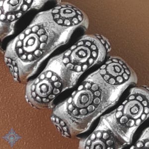 以下が含まれることがあります： 繰り返し花柄模様が施されたシルバーブレスレットのクローズアップ。円筒形のビーズは少し古びた外観です。背景は温かみのある茶色です。