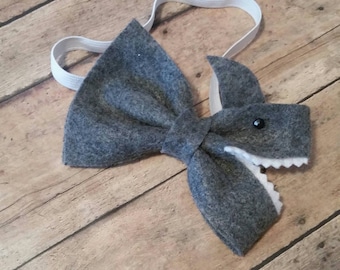 May include: A gray felt bow tie with a shark design. The bow tie has a central knot and a shark head with a black eye and white teeth. An elastic band is attached for wear.