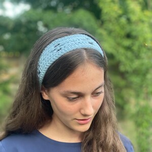 Puede incluir: Una diadema de ganchillo azul claro. La diadema se lleva en la cabeza. El diseño de ganchillo es visible. La persona de la imagen lleva una camiseta azul. El fondo está desenfocado.