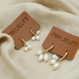 May include: Gold-tone hoop earrings with clusters of white pearls, displayed on brown leatherette cards. The cards are embossed with the word "MIKUKUMI". The earrings are set against a cream-colored fabric background.