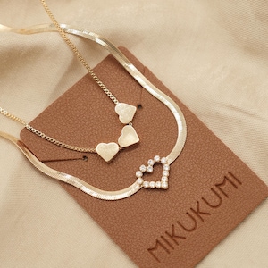 May include: Gold-toned necklaces with heart pendants. One necklace features a heart pendant encrusted with small, clear stones. The other necklace has three heart pendants. The necklaces are displayed on a brown card with the brand name "MIKUKUMI".