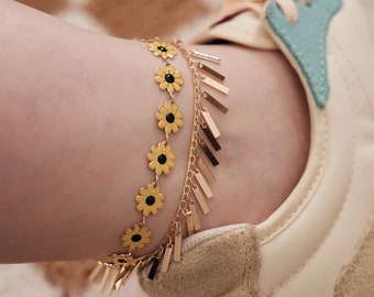 May include: Gold-toned anklet featuring daisy-shaped charms with black centers, and dangling rectangular accents. The anklet is worn on an ankle next to a beige and blue sneaker, set against a patterned background.