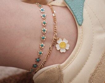 May include: Two gold anklets with a charm. One anklet has a chain with a small white flower charm with a yellow smiley face. The other anklet has a chain with small turquoise flowers.