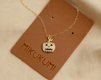 May include: A gold chain necklace with a small, sparkly pumpkin pendant. The pendant has a face with a carved out smile. The necklace is displayed on a brown leather tag with the word "MIKUKUMI" printed on it.