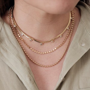 May include: A layered gold necklace with a delicate chain and a thicker chain with small round beads. The top chain has small clear stones hanging from it.