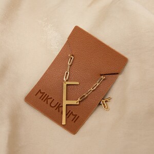 May include: A gold chain necklace with a gold letter 'F' pendant. The necklace is displayed on a brown leather pouch with the text 'MIKUKIMI' printed on it.