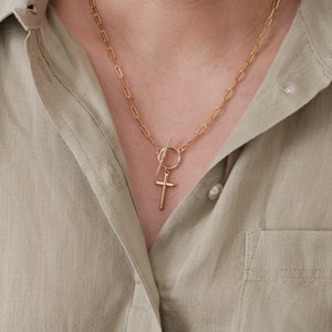 May include: A gold chain necklace with a toggle clasp and a small cross pendant. The necklace is worn by a person wearing a light green shirt.