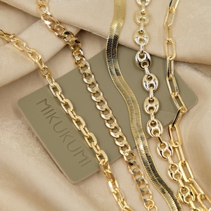May include: Four gold chains of different styles and thicknesses lay on a beige fabric surface. The chains are arranged in a criss-cross pattern. A small, rectangular, light brown card with the text "MIKUKUMI" is visible in the background.
