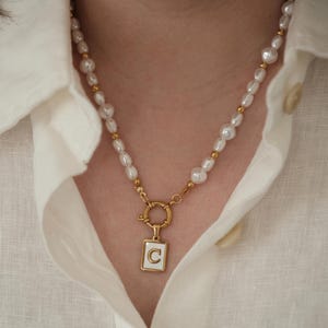 May include: A gold necklace with a white pearl chain and a gold pendant with the letter "C" in a white square.