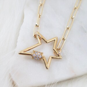 May include: A gold-colored necklace featuring a star-shaped pendant with a small, cylindrical clasp adorned with clear crystals. The chain is made of rectangular links. The necklace is displayed on a white, fluffy surface.