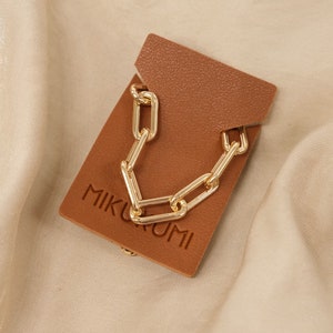 Pode incluir: Um colar de corrente de clipe de papel grosso dourado em um cart&atilde;o de couro marrom com a palavra "MIKKUMI" impressa nele. "Chunky Paperclip Chain" est&aacute; escrito abaixo do cart&atilde;o.