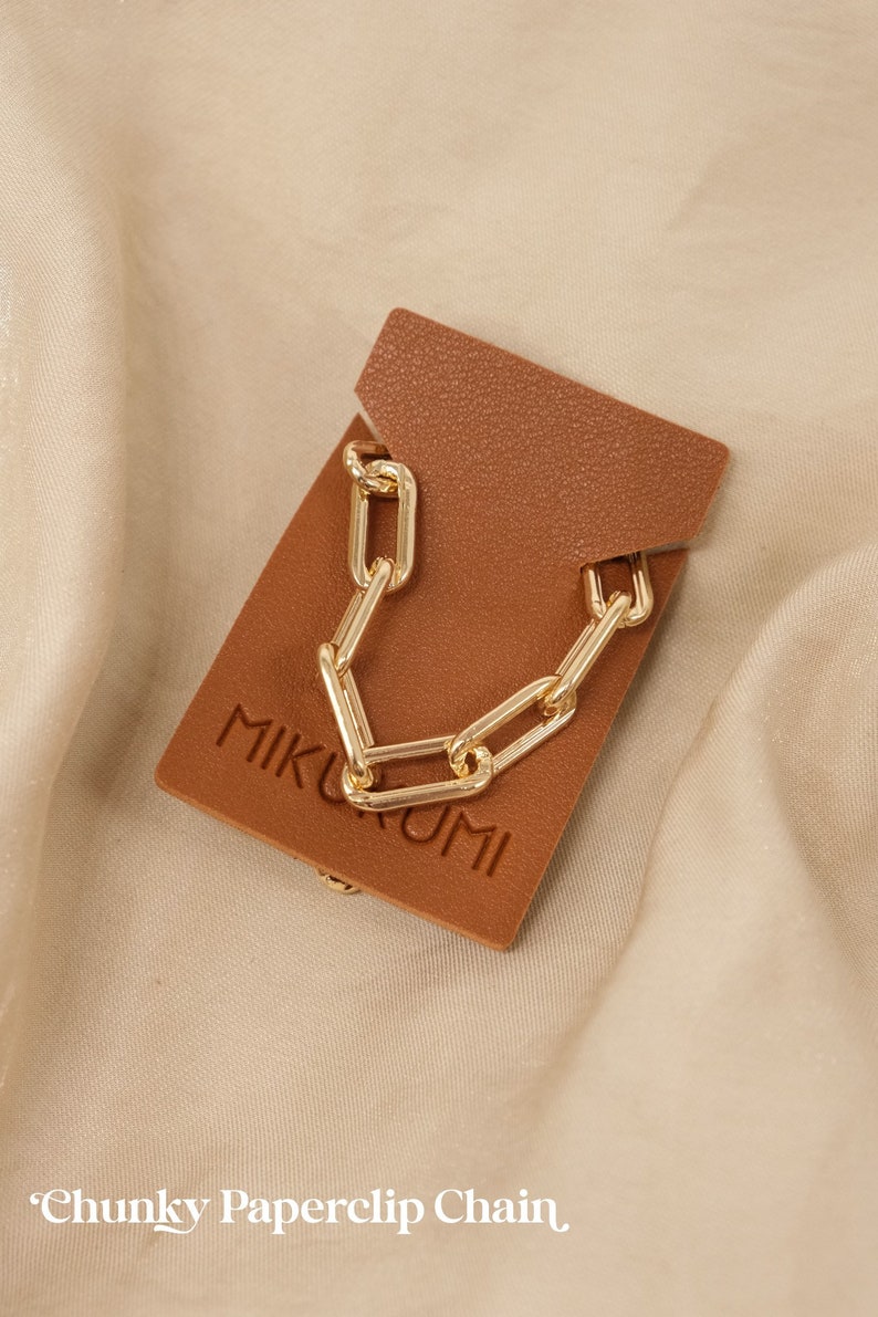 Pode incluir: Um colar de corrente de clipe de papel grosso dourado em um cart&atilde;o de couro marrom com a palavra "MIKKUMI" impressa nele. "Chunky Paperclip Chain" est&aacute; escrito abaixo do cart&atilde;o.