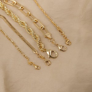 Pode incluir: Cinco correntes de ouro de diferentes estilos e comprimentos, incluindo uma corrente de corda, uma corrente de curb, uma corrente de caixa e uma corrente de cobra. As correntes est&atilde;o dispostas sobre um fundo de tecido bege.