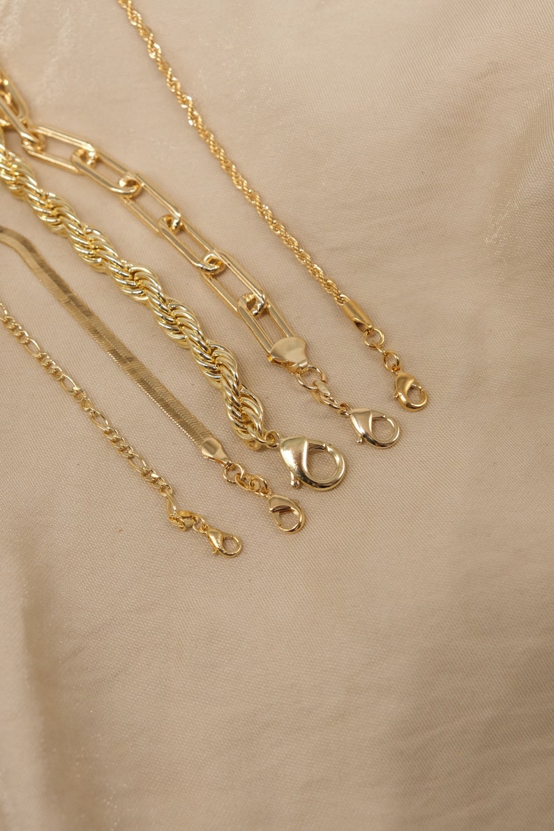 Pode incluir: Cinco correntes de ouro de diferentes estilos e comprimentos, incluindo uma corrente de corda, uma corrente de curb, uma corrente de caixa e uma corrente de cobra. As correntes est&atilde;o dispostas sobre um fundo de tecido bege.