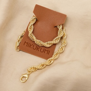 Pode incluir: Um colar de corrente de corda grossa de ouro com um fecho de lagosta. A corrente &eacute; exibida em uma etiqueta de couro marrom com a palavra "MIKUKUMI" impressa nela. O texto "Chunky Rope Chain" est&aacute; escrito abaixo da imagem.