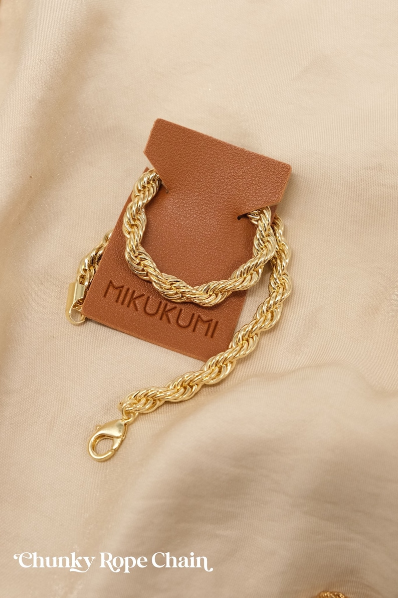 Pode incluir: Um colar de corrente de corda grossa de ouro com um fecho de lagosta. A corrente &eacute; exibida em uma etiqueta de couro marrom com a palavra "MIKUKUMI" impressa nela. O texto "Chunky Rope Chain" est&aacute; escrito abaixo da imagem.