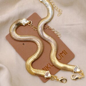 May include: Two gold-toned, snake chain necklaces with a textured, ribbed design. The necklaces are displayed on a brown tag with the text "IKU UMI". Each necklace has a lobster clasp closure and an extender chain.