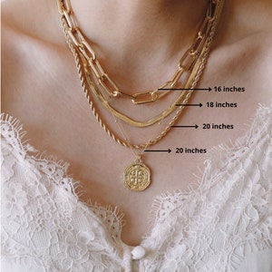 Pode incluir: Quatro colares de corrente de ouro sobrepostos no pesco&ccedil;o de uma pessoa. As correntes medem 40, 45, 50 e 50 cent&iacute;metros de comprimento. A corrente inferior tem um pingente de moeda de ouro com um desenho de cruz.