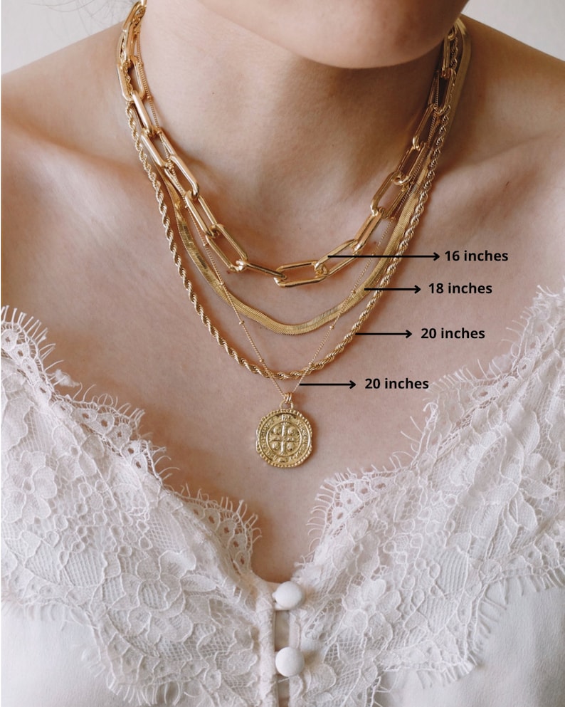 Pode incluir: Quatro colares de corrente de ouro sobrepostos no pesco&ccedil;o de uma pessoa. As correntes medem 40, 45, 50 e 50 cent&iacute;metros de comprimento. A corrente inferior tem um pingente de moeda de ouro com um desenho de cruz.