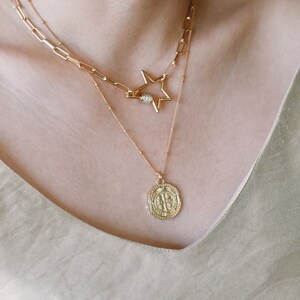 May include: A layered gold necklace set. The top layer features a star-shaped pendant with a central gem. The second layer is a chain with a coin pendant. The necklaces are displayed on a person's chest, against a neutral background.
