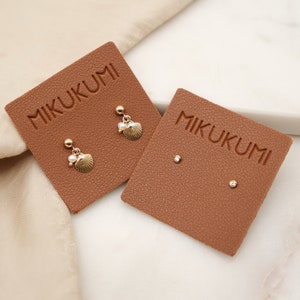 May include: Gold-toned seashell and pearl earrings displayed on a brown card with the brand name MIKUKUMI. A pair of stud earrings with a small diamond and a gold ball stud are also shown. The background is a marble surface.