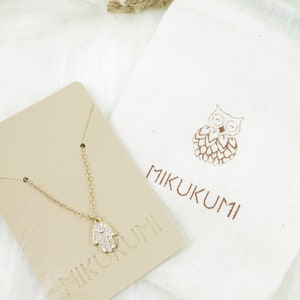 May include: A gold-colored necklace with a Hamsa hand pendant, encrusted with small, clear crystals. The necklace rests on a beige card with the brand name "MIKUKUMI" printed on it. A white drawstring bag with a brown owl design and the brand name is also present.