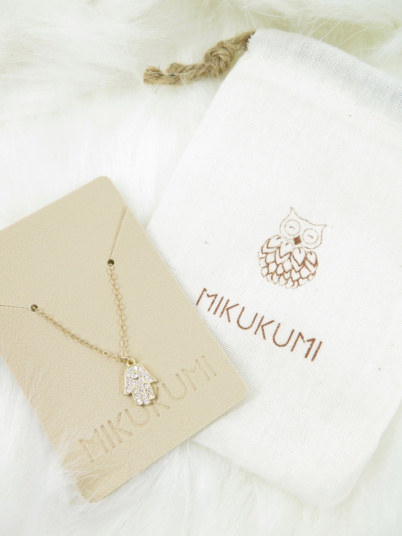 May include: A gold-colored necklace with a Hamsa hand pendant, encrusted with small, clear crystals. The necklace rests on a beige card with the brand name "MIKUKUMI" printed on it. A white drawstring bag with a brown owl design and the brand name is also present.