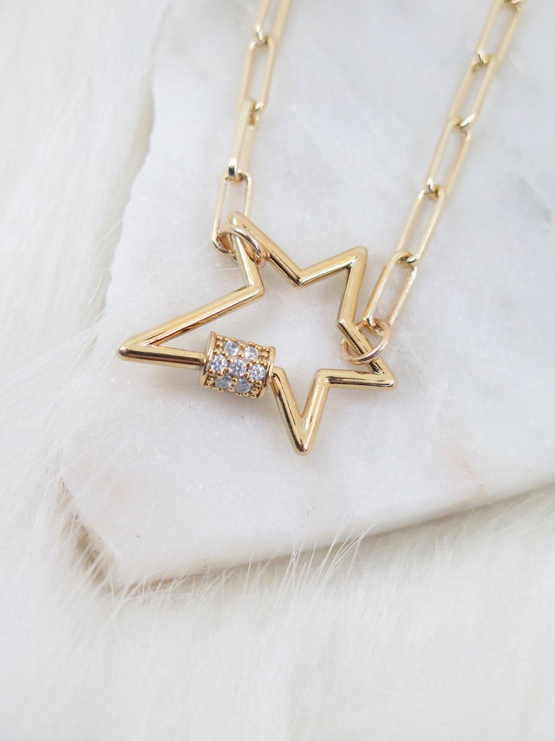 May include: A gold-colored necklace featuring a star-shaped pendant with a small, cylindrical clasp adorned with clear crystals. The chain is made of rectangular links. The necklace is displayed on a white, fluffy surface.