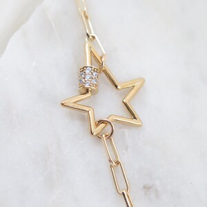 May include: Gold-colored necklace featuring a star pendant. The star is hollow with a five-point design. The necklace includes a cylindrical clasp adorned with small, clear gemstones and a chain link design.