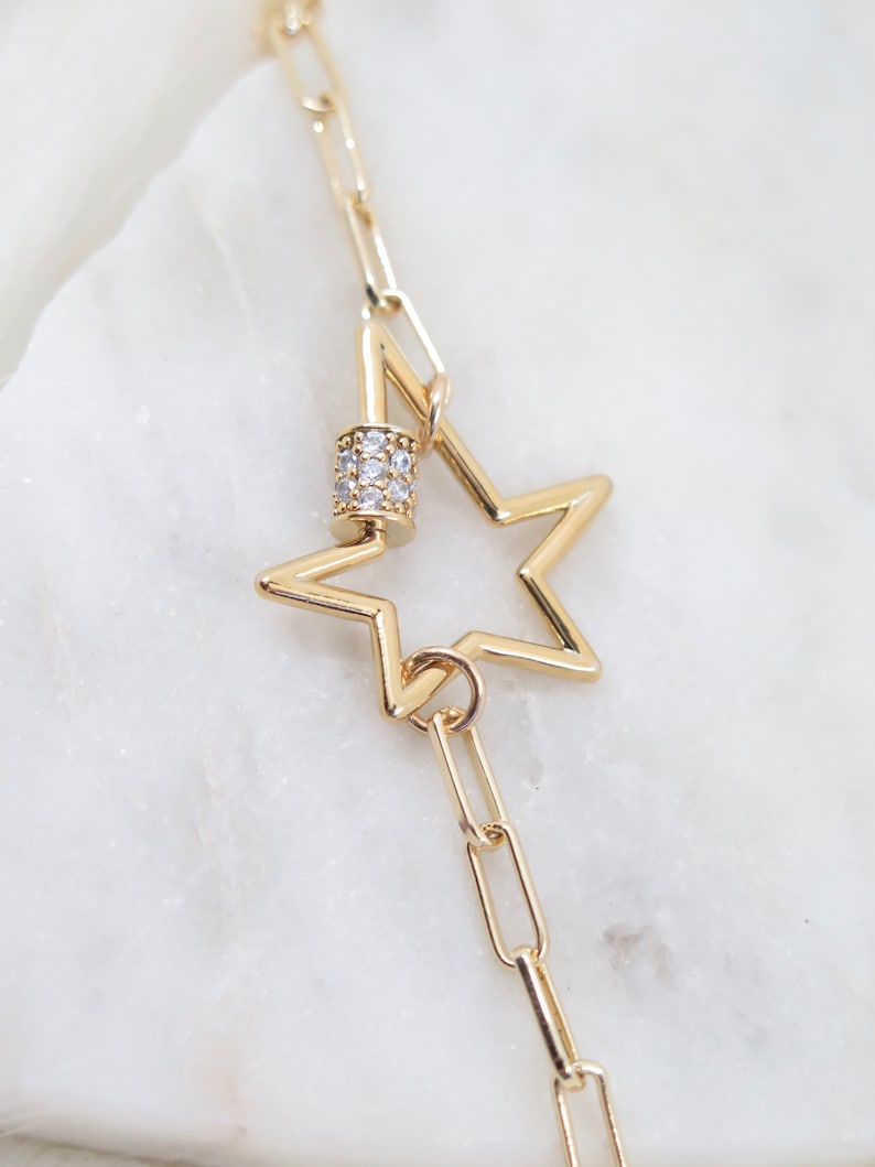 May include: Gold-colored necklace featuring a star pendant. The star is hollow with a five-point design. The necklace includes a cylindrical clasp adorned with small, clear gemstones and a chain link design.
