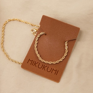 Pode incluir: Um colar de corrente de corda dourada em uma etiqueta de couro marrom com o texto "MIKUKUMI" impresso nela. A etiqueta est&aacute; em um fundo de tecido bege. Corrente de corda.