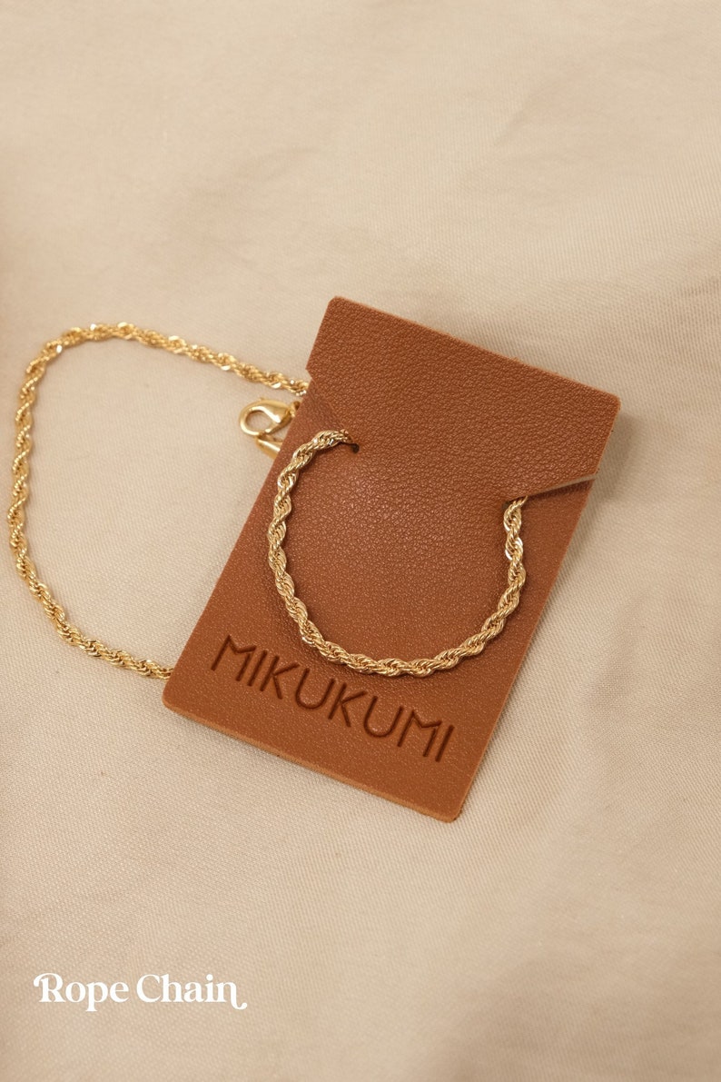 Pode incluir: Um colar de corrente de corda dourada em uma etiqueta de couro marrom com o texto "MIKUKUMI" impresso nela. A etiqueta est&aacute; em um fundo de tecido bege. Corrente de corda.