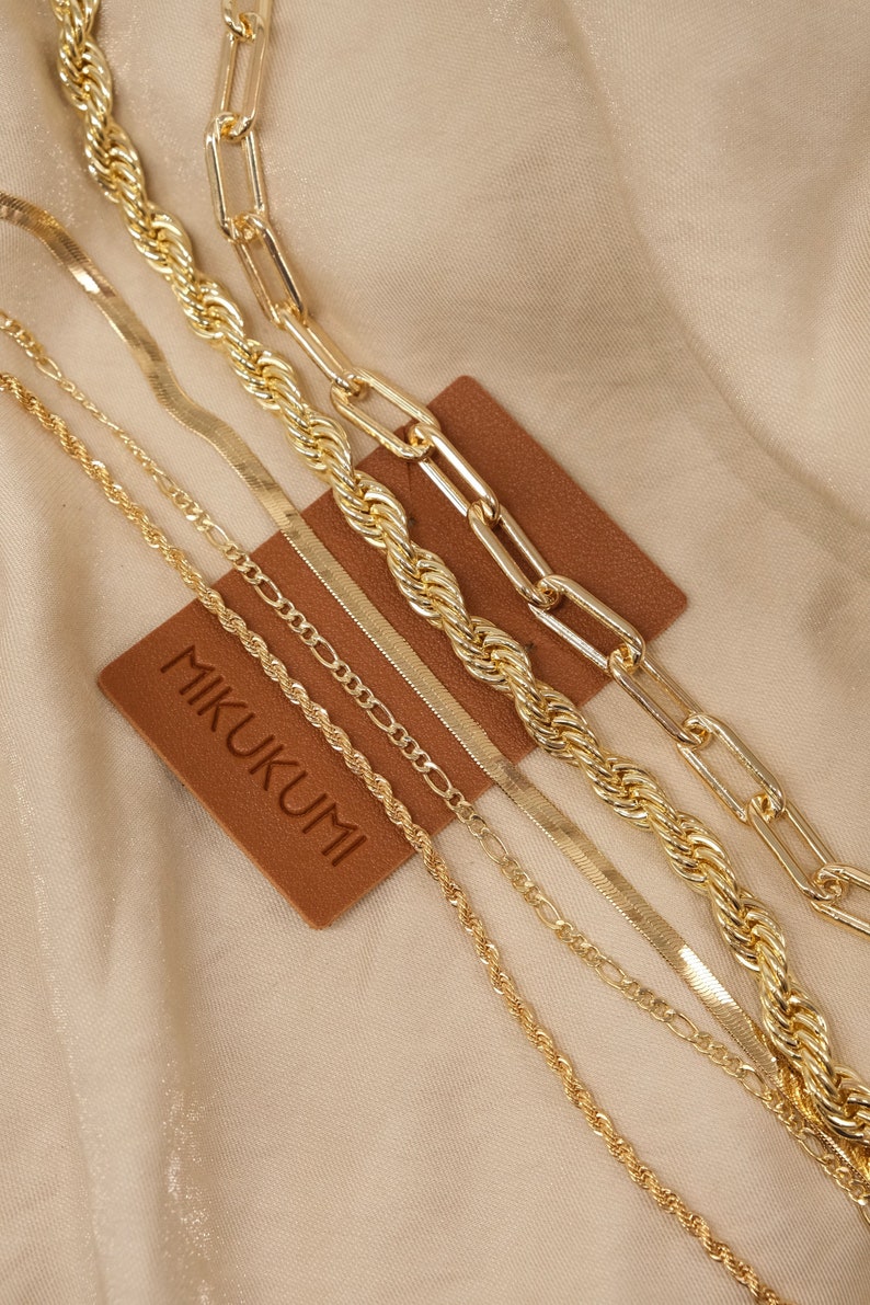 Pode incluir: Um conjunto de cinco correntes douradas de diferentes estilos e espessuras, incluindo uma corrente de corda, uma corrente de curb, uma corrente de caixa e uma corrente de espinha de peixe. As correntes est&atilde;o dispostas em uma etiqueta de couro marrom com a palavra "MIKUKUMI" impressa nela.
