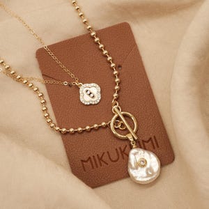 May include: A gold chain necklace with a small, round pendant with clear stones and a larger, irregular shaped pendant with a pearl and a clear stone. The necklace is on a brown leather background with the word "MIKUKIMI" printed on it.
