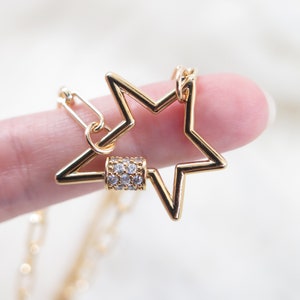 May include: A gold-colored star-shaped pendant necklace. The star has a hollow design and a small, cylindrical clasp adorned with clear crystals. The necklace chain is also gold and visible in the image.