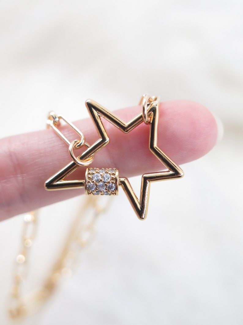 May include: A gold-colored star-shaped pendant necklace. The star has a hollow design and a small, cylindrical clasp adorned with clear crystals. The necklace chain is also gold and visible in the image.