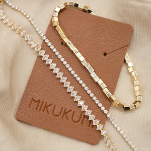 May include: Three gold-toned bracelets with clear crystal accents and a brown tag. One bracelet features square links, while the others have a delicate chain design. The tag displays the brand name "MIKUKU".