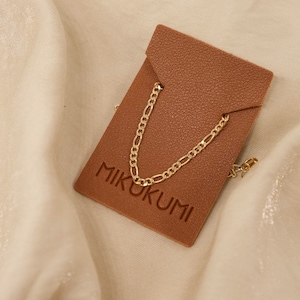 Pode incluir: Um colar de corrente figaro dourado em uma etiqueta de couro marrom com a palavra "MIKUKUMI" gravada.