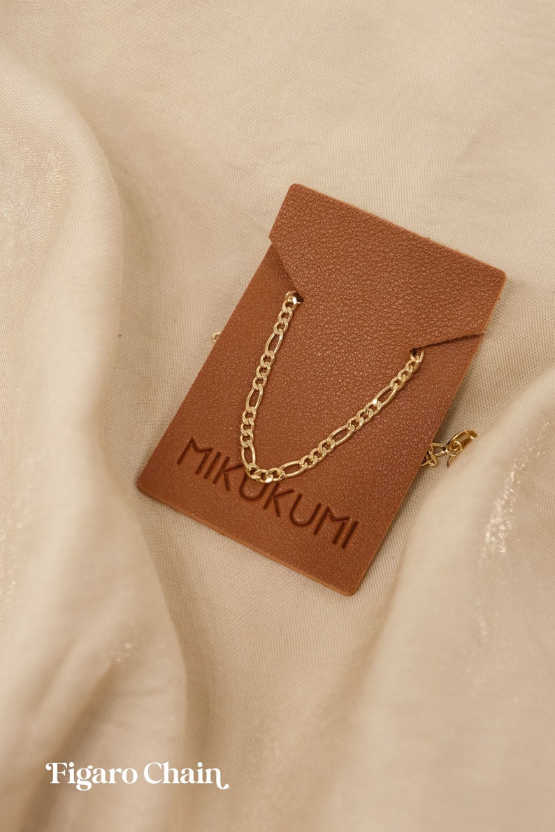 Pode incluir: Um colar de corrente figaro dourado em uma etiqueta de couro marrom com a palavra "MIKUKUMI" gravada.