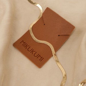 Pode incluir: Um colar de corrente de cobra ou espinha de peixe dourado em uma etiqueta de couro marrom com a palavra "MIKUKUMI" impressa nela.