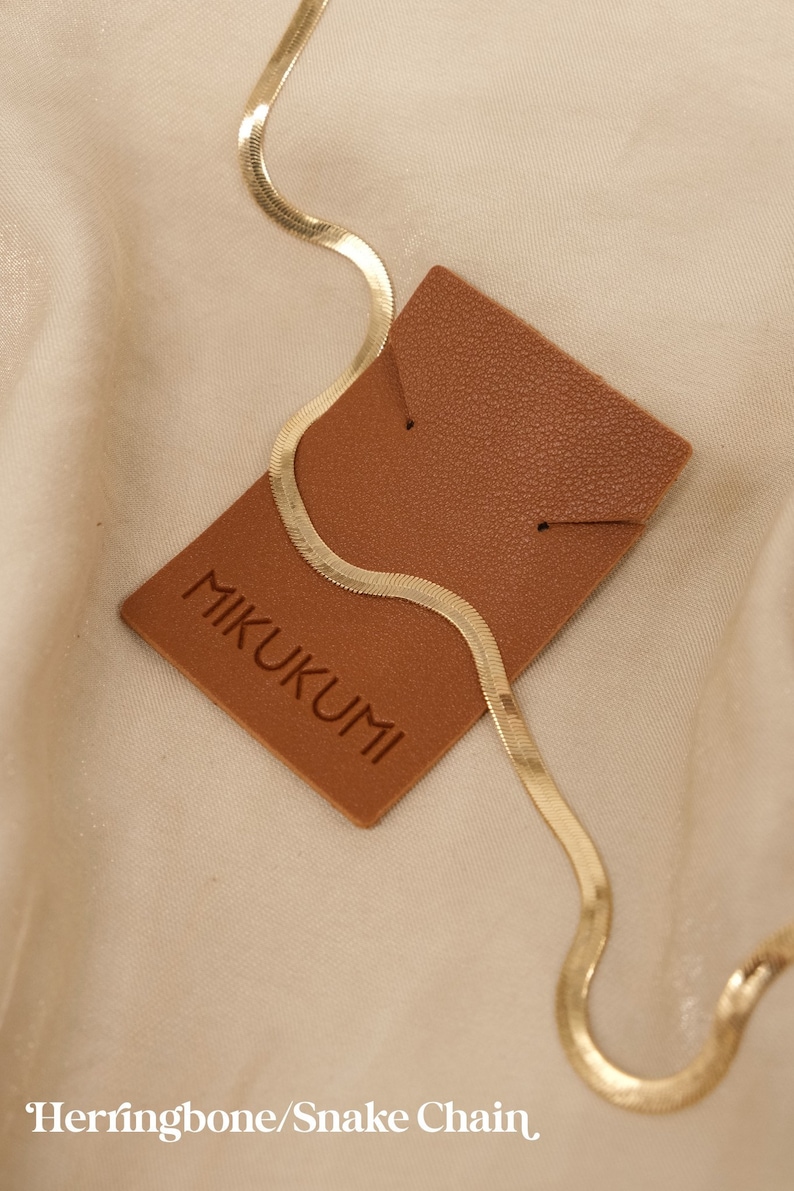 Pode incluir: Um colar de corrente de cobra ou espinha de peixe dourado em uma etiqueta de couro marrom com a palavra "MIKUKUMI" impressa nela.